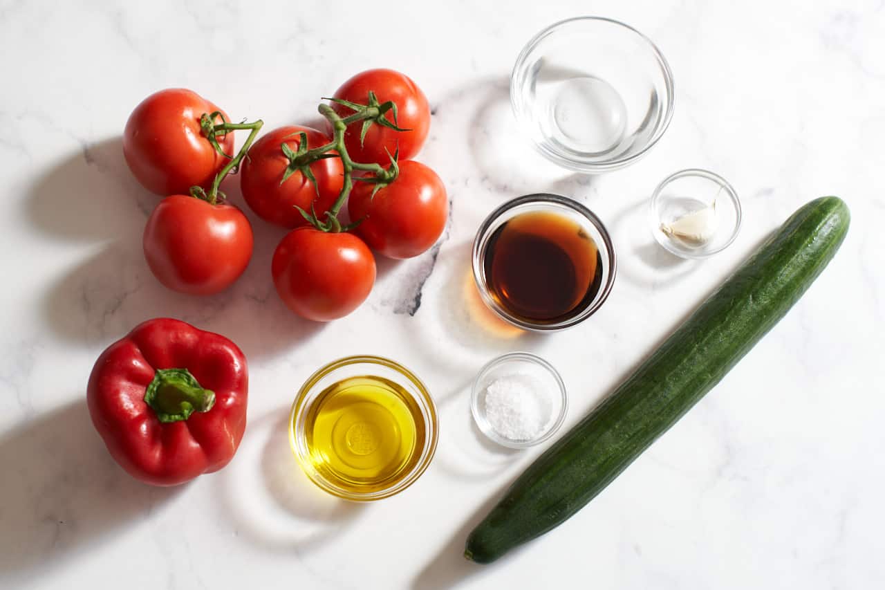Tomatoes, a bell pepper, an English cucumber, and small glass bowls of olive oil, sherry vinegar, salt, garlic and water.