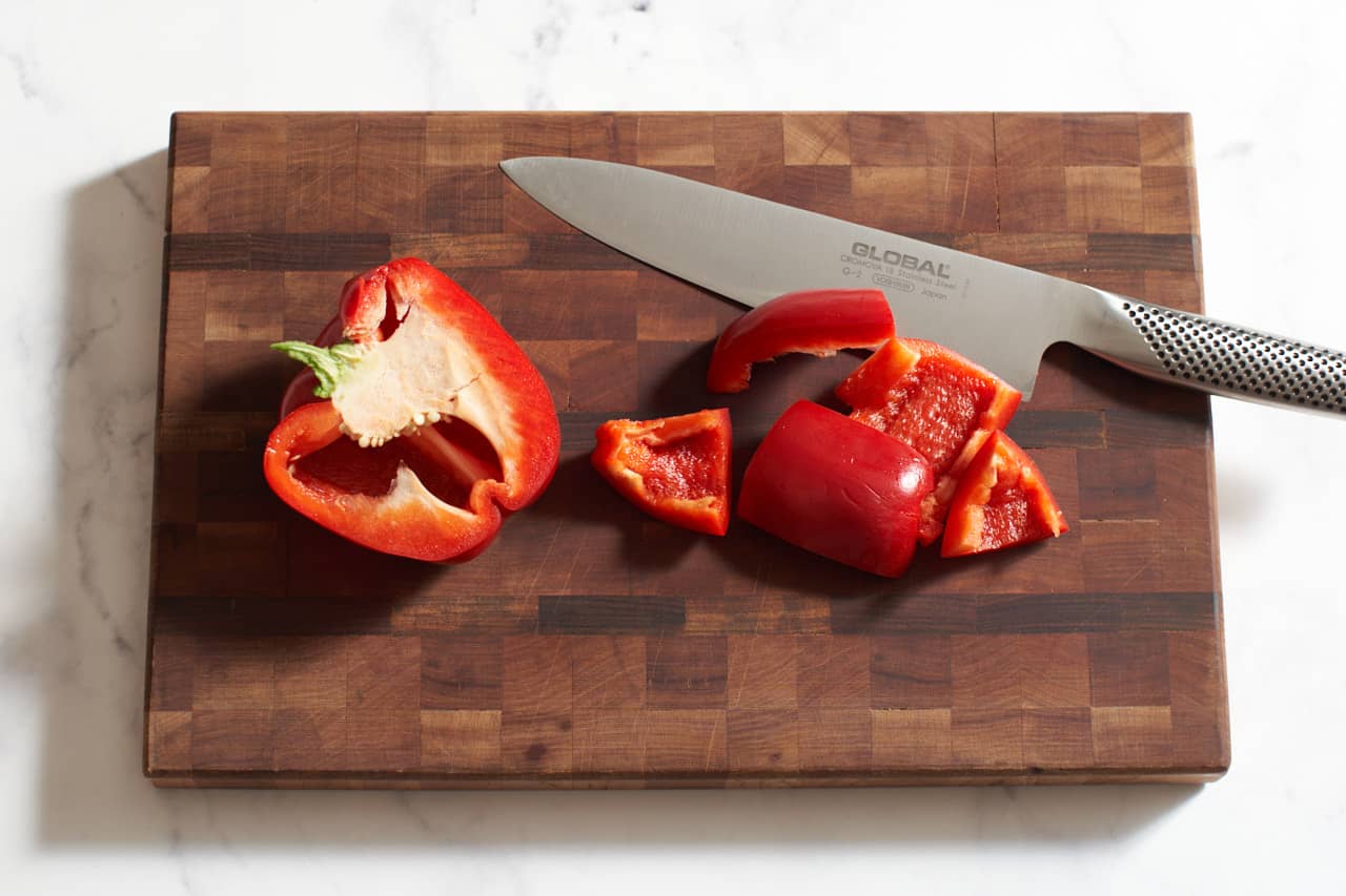 A knife on a wooden cutting board with a red pepper that has been halved and cut into pieces.