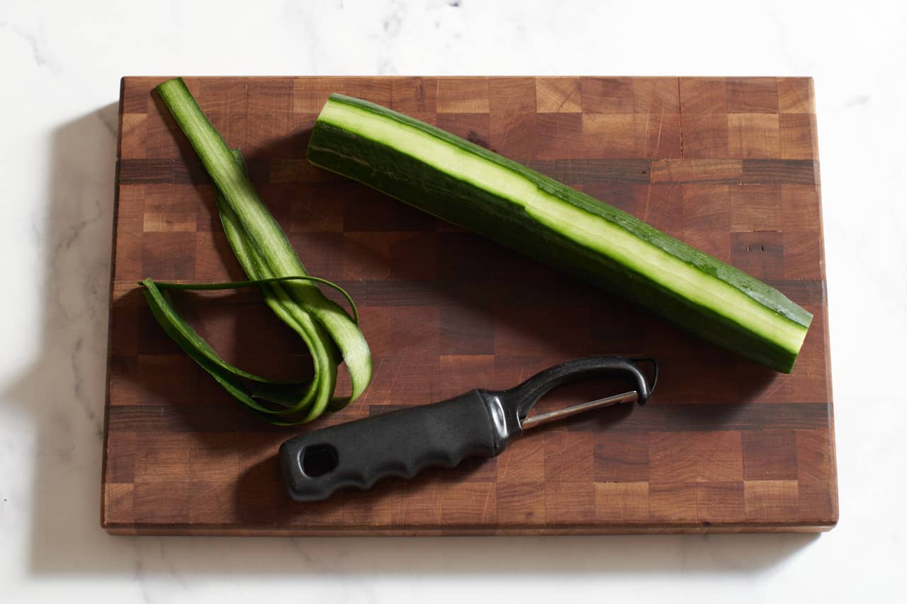 A vegetable peeler on a wooden cutting board with a cucumber that has strips of peel removed.
