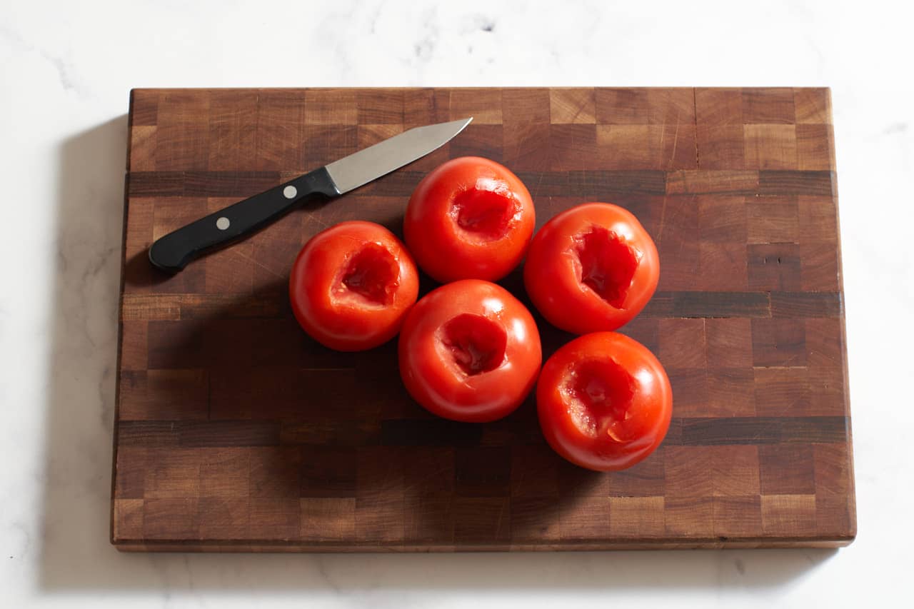 A paring knife on a wooden cutting board next to five tomatoes that have been cored.