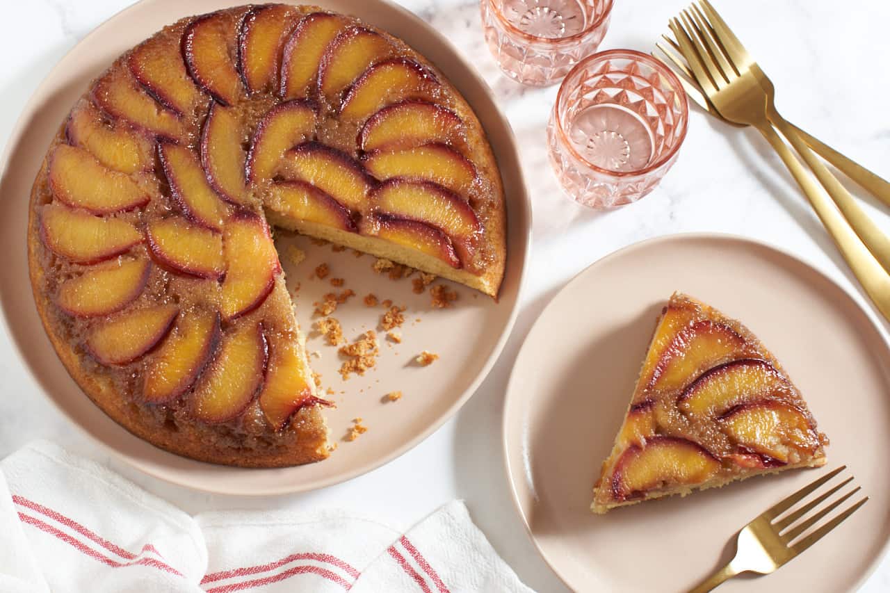 A plum upside down cake with a slice cut out of it, next to a pink plate with a slice of cake and a gold fork on it. Gold forks, two pink glasses, and a red and white striped towel surround the cake.