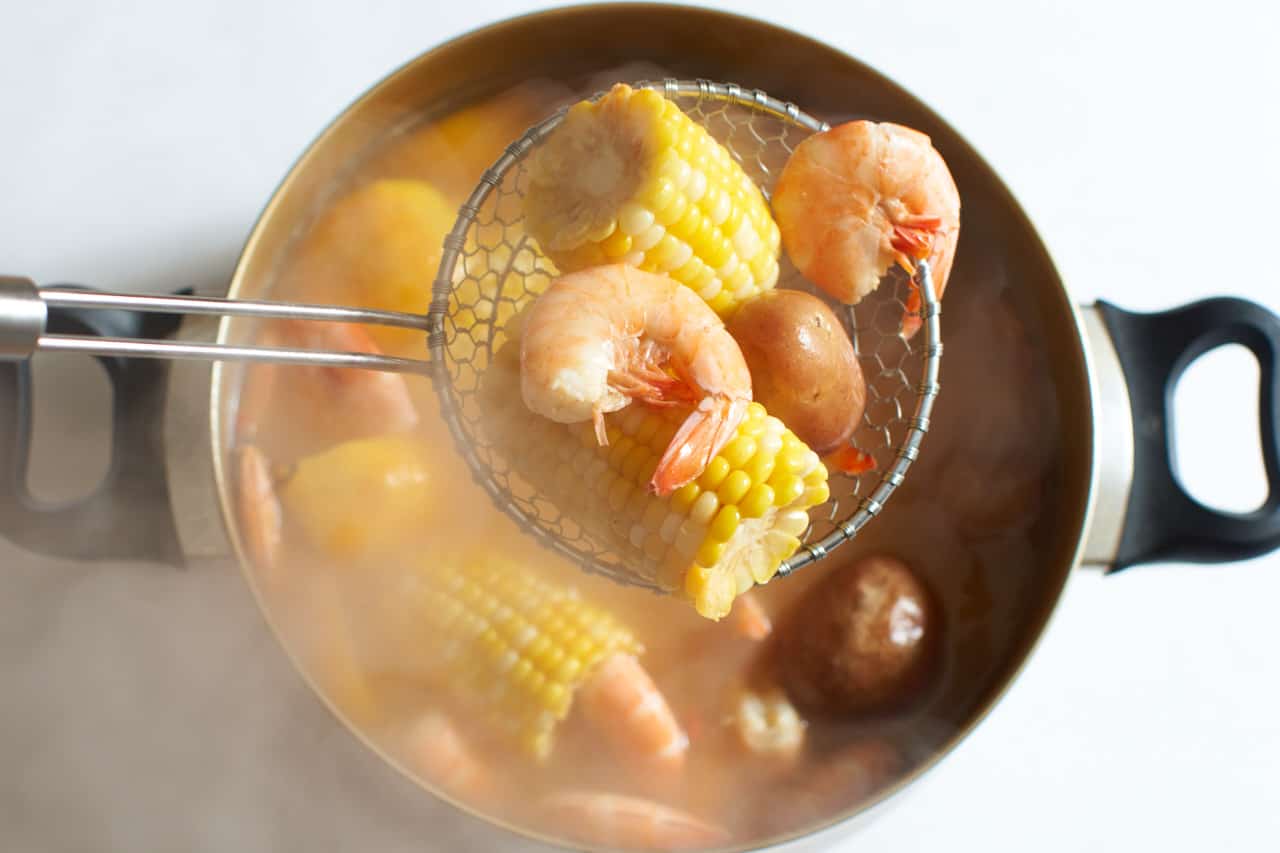 A strainer full of steaming shrimp, potatoes, and corn being lifted out of a pot of low country boil..