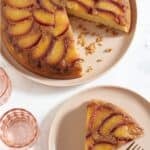 A caramelized plum upside down cake with a slice cut out of it above a pink plate that has a slice of cake on it.