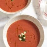 Two bowls of gazpacho topped with chopped cucumber with a red and white striped towel.