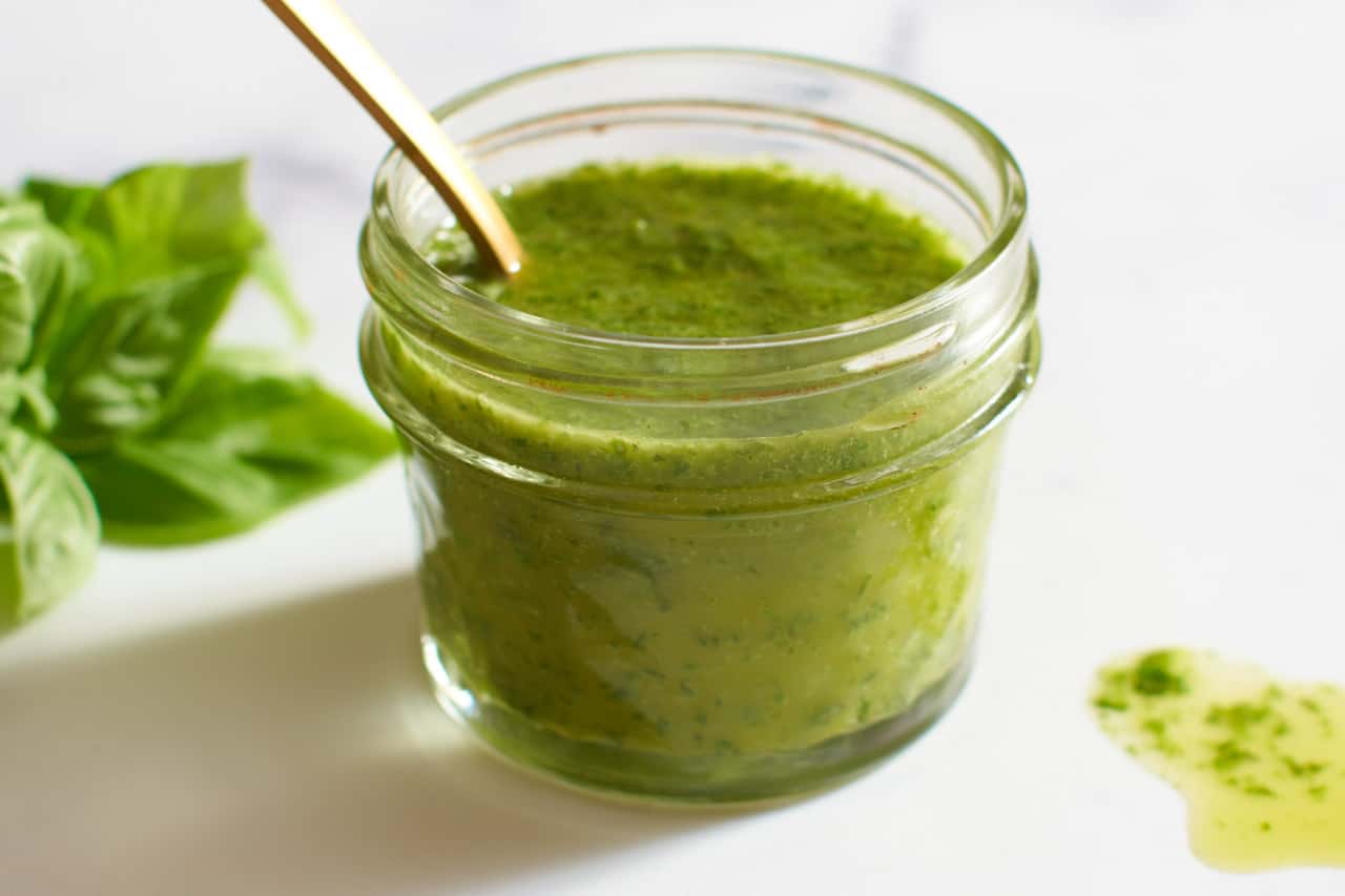 A small jar of basil vinaigrette with a gold spoon in it. A bunch of fresh basil is in the background, some spilled vinaigrette is in the foreground.