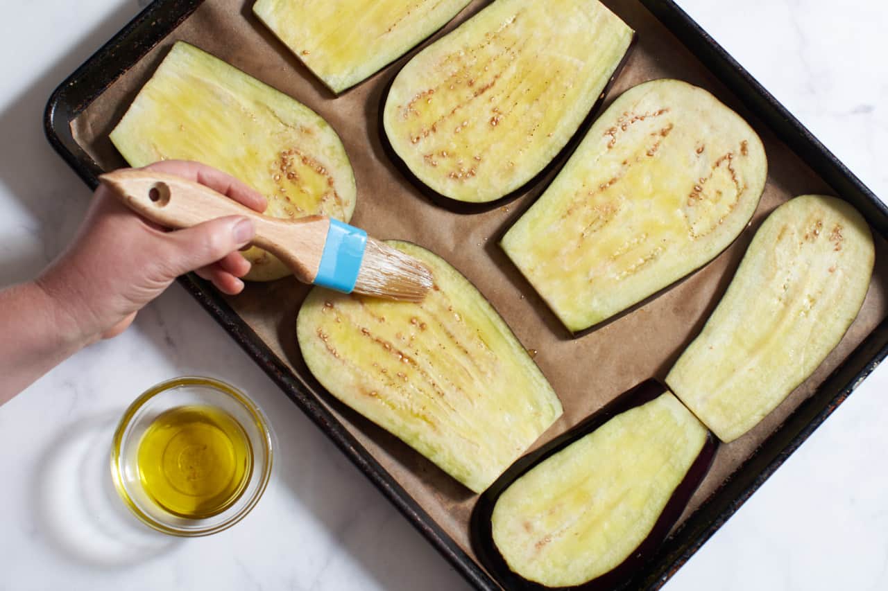 A woman's hand holding a pastry brush, brushing olive oil onto eggplant slices.