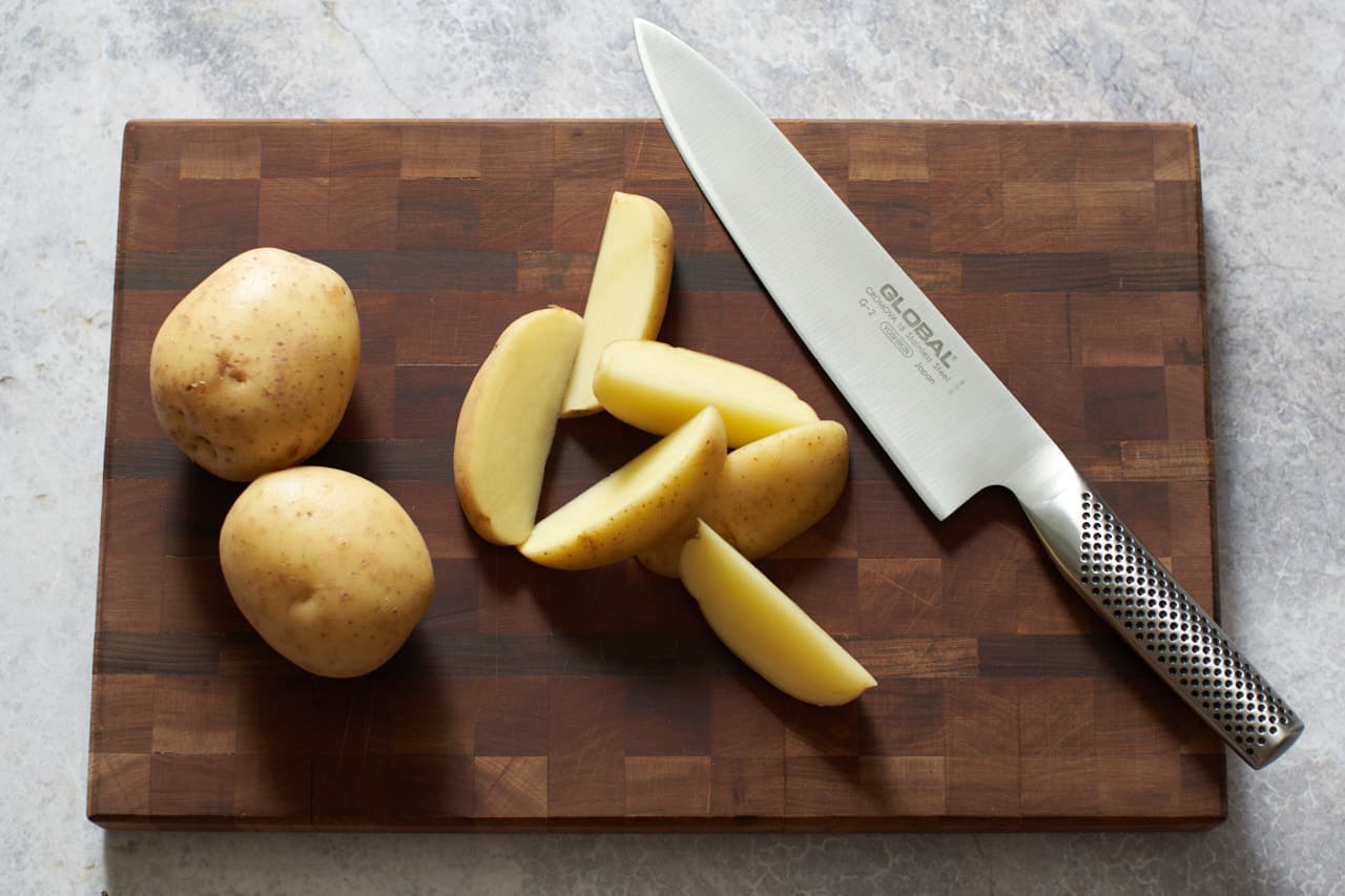 A knife on a wooden cutting board with potatoes that have been cut into wedges.