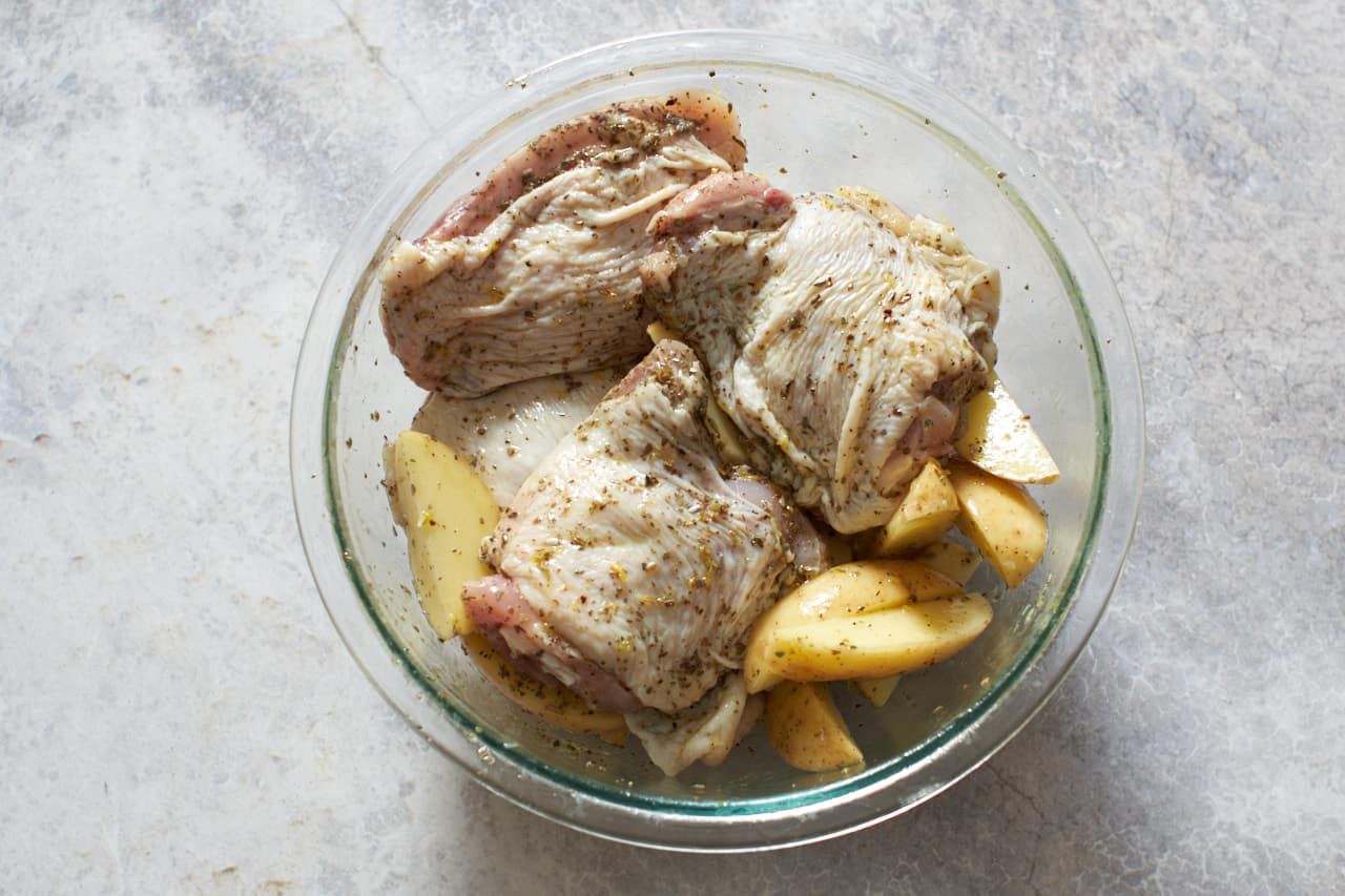 A bowl of chicken thighs and potatoes marinating in olive oil and spices.