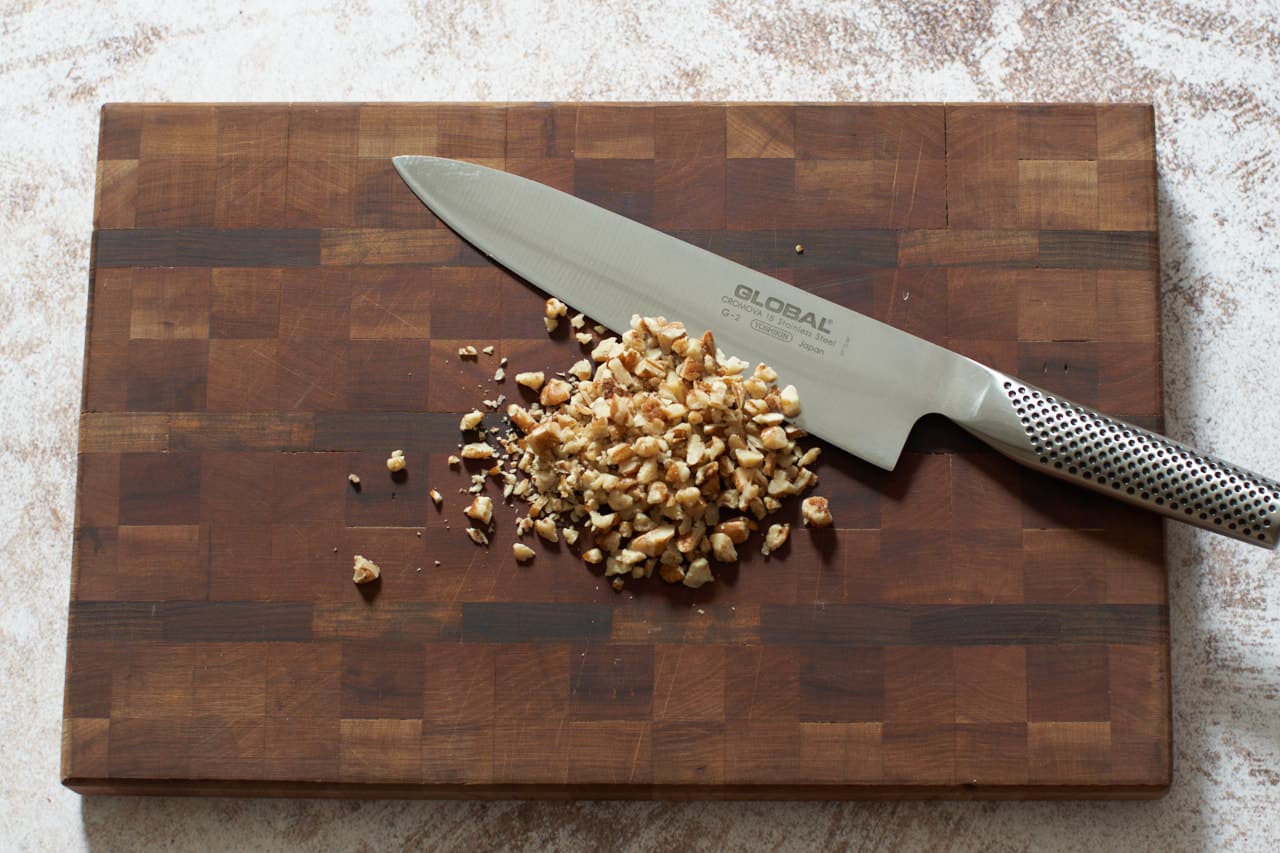 A knife on a wooden cutting board with chopped pecans.