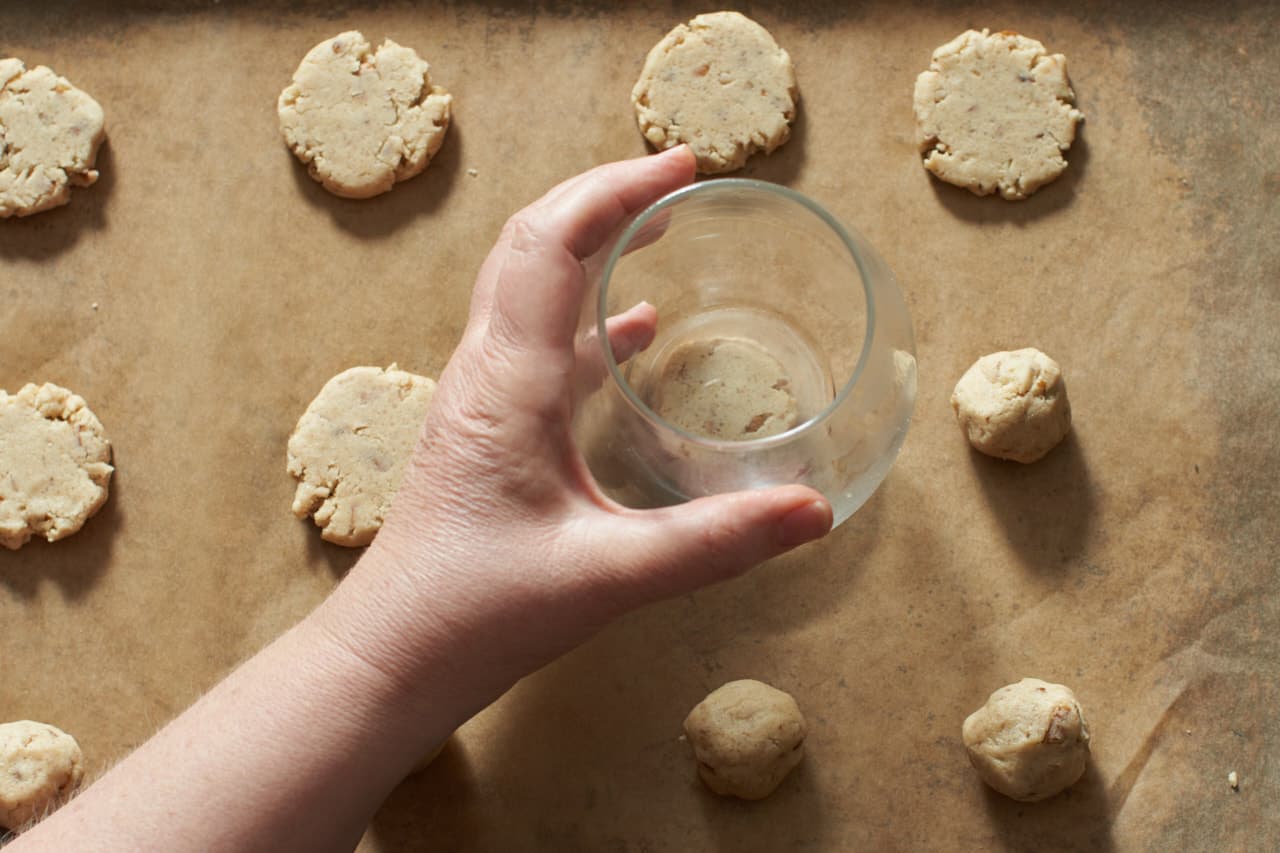 A woman's hand holding a wine glass that is pressing down on balls of pecan sandies cookie dough to flatten them.