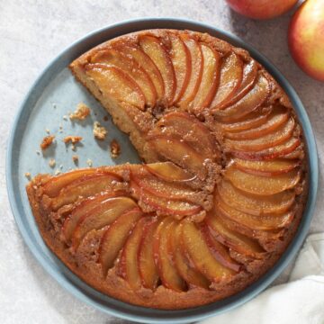 A caramel apple upside down cake with a sliced removed on a blue plate. Two apples are in the upper right hand corner.