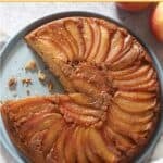 A caramel apple upside down cake with a sliced removed on a blue plate, two apples are in the upper right hand corner.