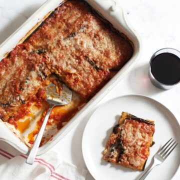 A casserole dish of vegetarian eggplant lasagna with a spatula in it next to a glass of red wine, and a plate with a slice of lasagna on it.