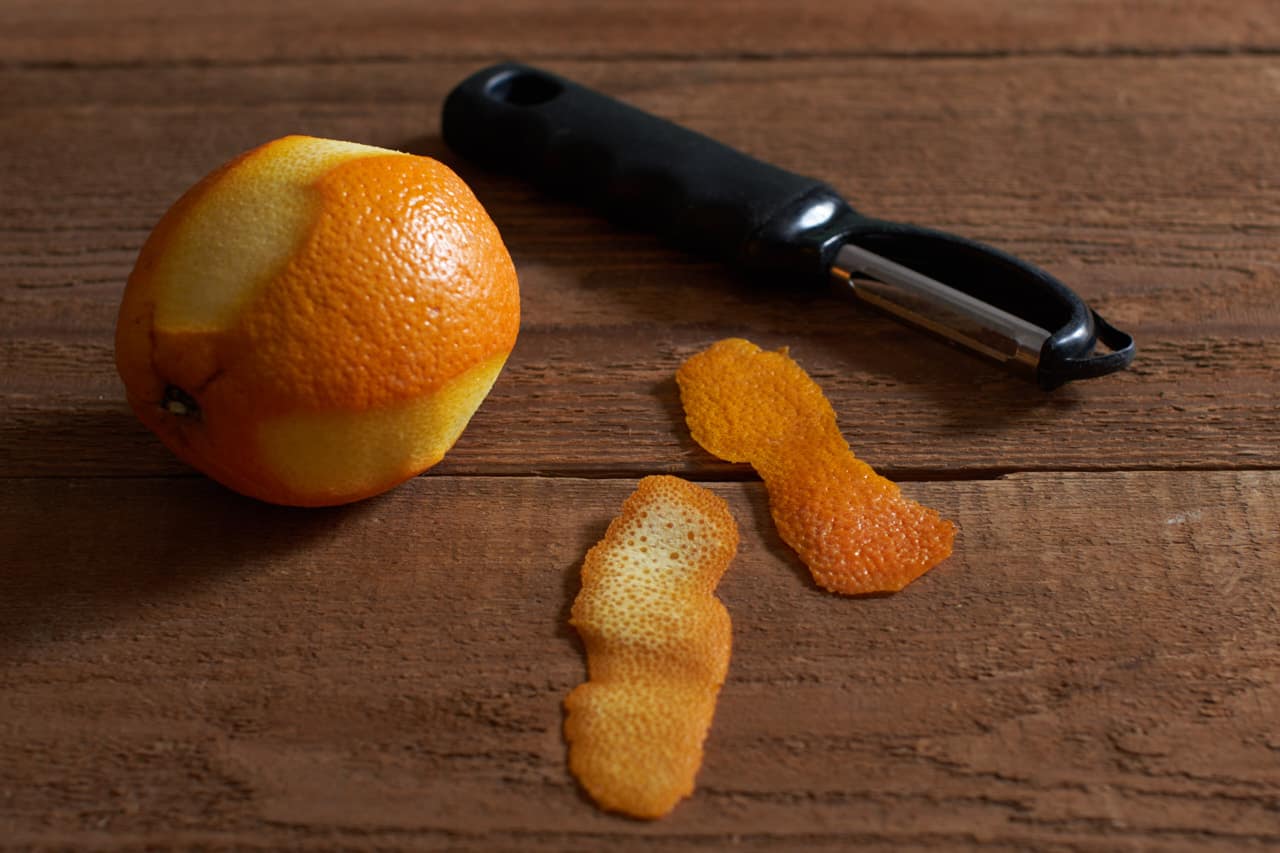 A vegetable peeler next to an orange and two strips of orange zest.