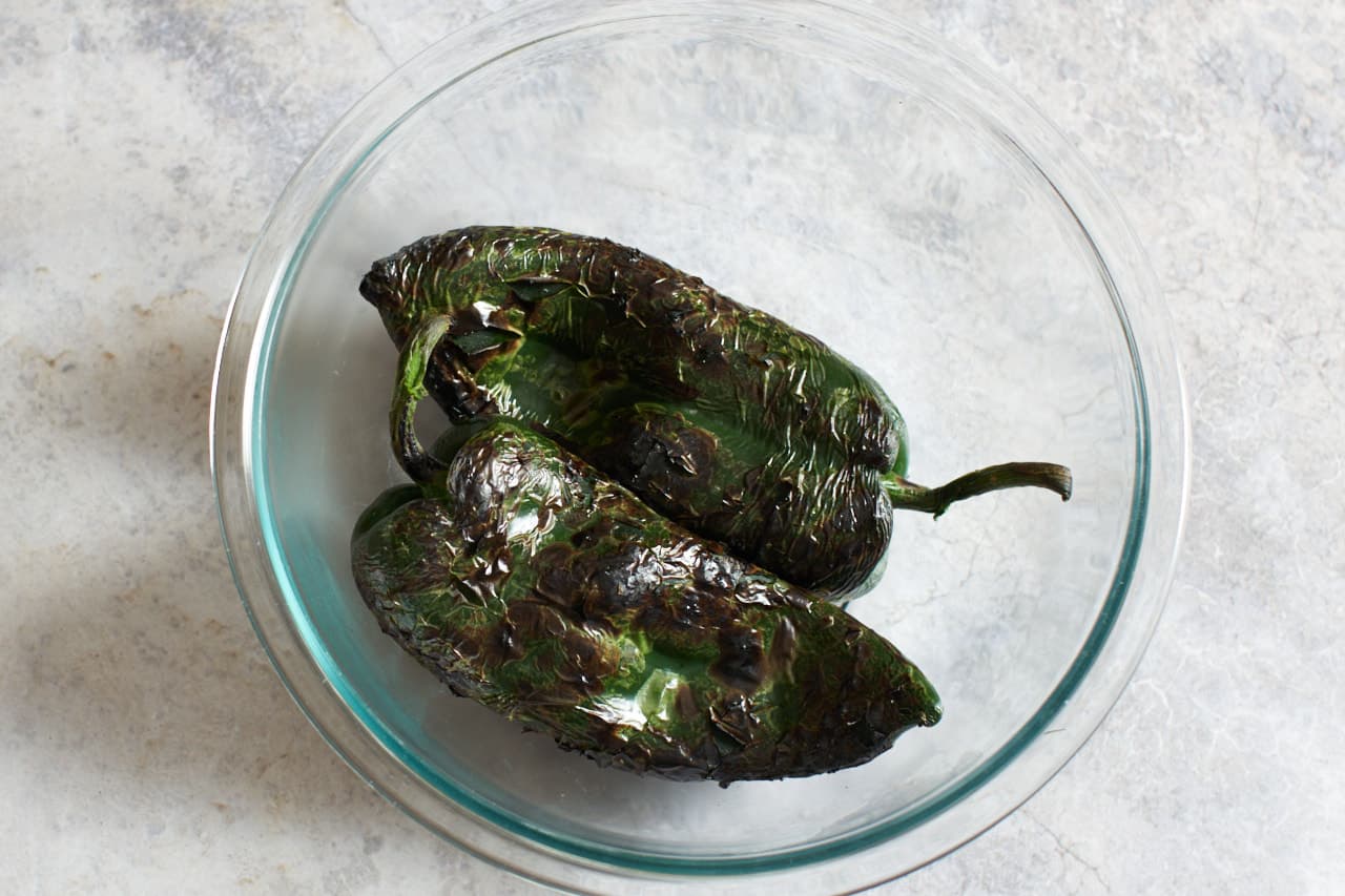 Two roasted poblano peppers in a glass bowl.
