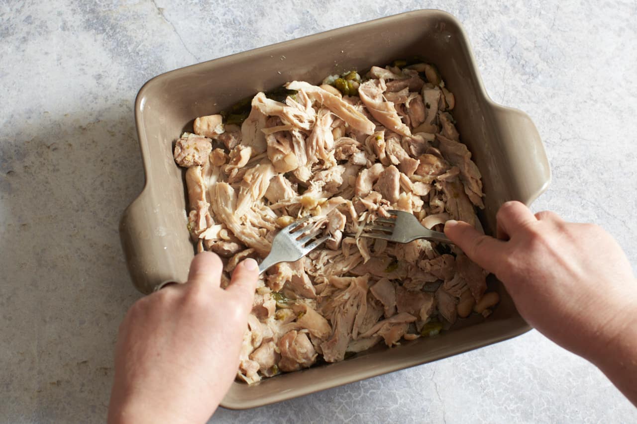 A woman's hands are shown pulling cooked chicken apart with two forks.
