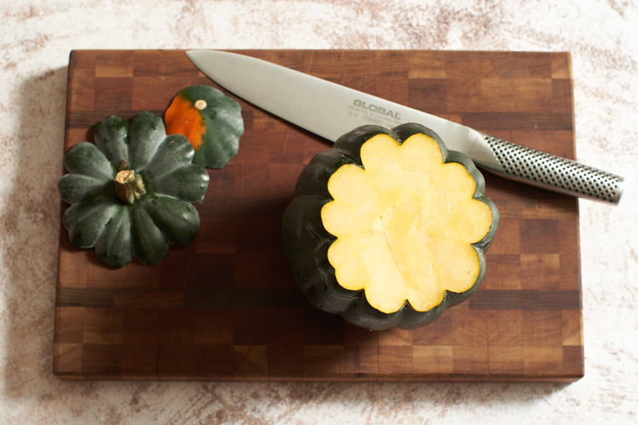 An acorn squash with the top and bottom sliced off on a wooden cutting board with a chef's knife.