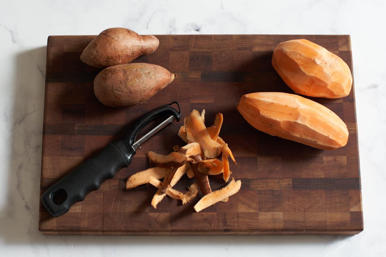Sweet potatoes and sweet potato peels on a wooden cutting board with a vegetable peeler.