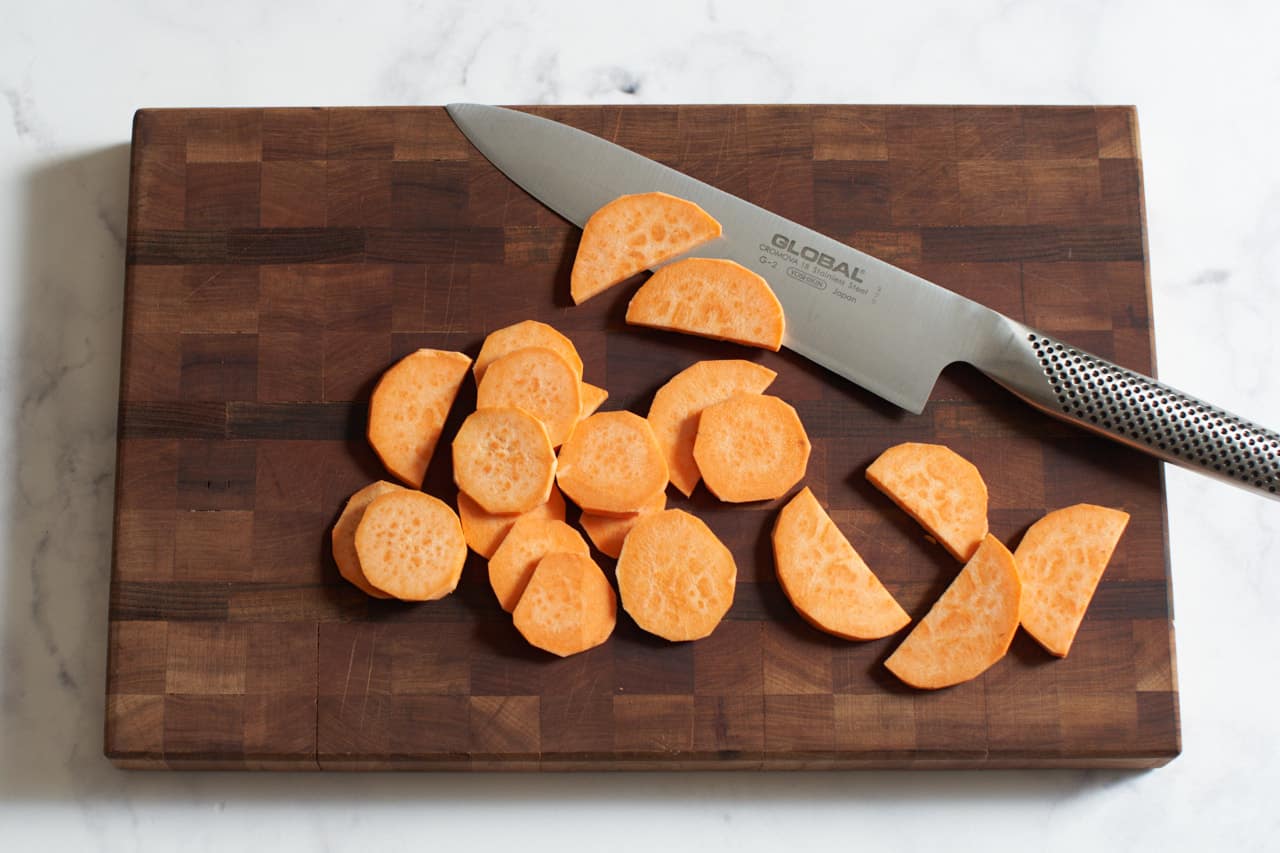 A chef's knife on a wooden cutting board with slices of sweet potatoes.