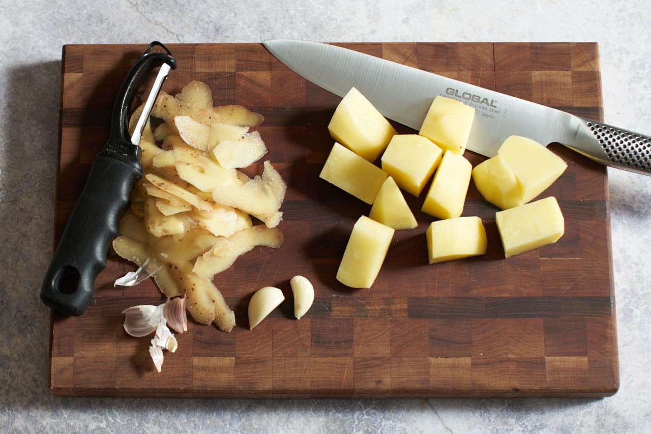 Cubed potatoes, garlic cloves, potato peels, a vegetable peeler, and a chef's knife on a cutting board.