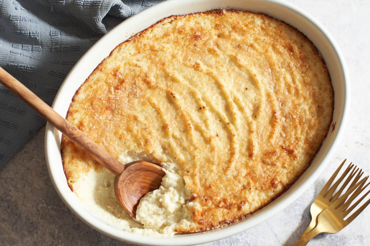 An oval gratin dish of goat cheese mashed potatoes with a wooden spoon in it. A blue towel is in the upper left corner, two gold forks are on the bottom right.