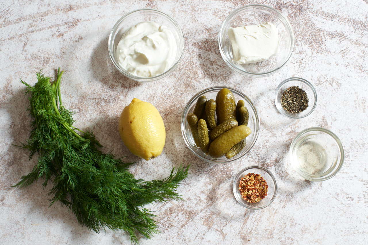 A lemon, fresh dill, and small bowls of pickles, sour cream, cream cheese, pickle brine, black pepper and crushed red pepper.
