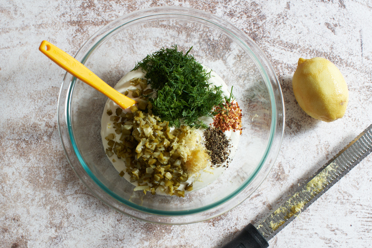 A bowl of cream cheese/sour cream mixture with pickles, dill, lemon zest, pepper, and crushed red pepper on top. A lemon and a grater are to the right.