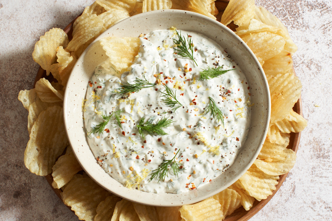 A bowl of dill pickle dip topped with fresh dill, pepper, and lemon zest, surrounded by ridged potato chips.
