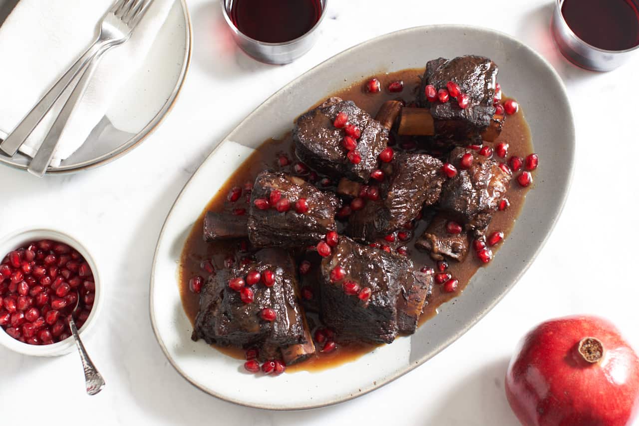 A plate of braised short ribs in pomegranate wine sauce topped with pomegranate seeds. Two glasses of wine, a fresh pomegranate, a bowl of pomegranate seeds and extra plates surround the platter.