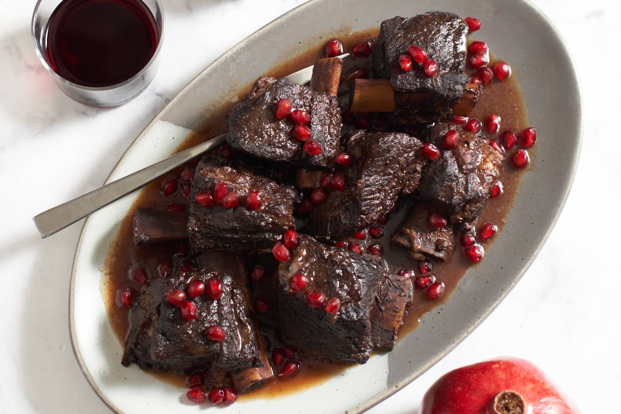 A platter of short ribs in a pomegranate wine sauce with pomegranate seeds on top with a serving fork on the left side of the plate. A glass of red wine is in the upper left and a pomegranate is in the lower right.