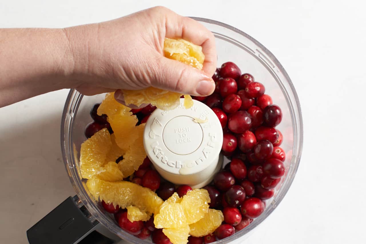 A woman's hand squeezing orange juice into a food processor with fresh cranberries and orange wedges in it.