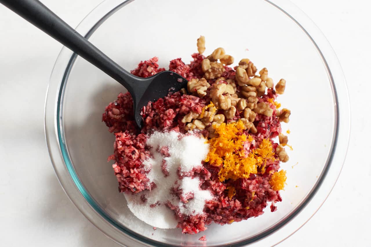 Chopped cranberries, orange zest, sugar and walnuts in a glass bowl with a black spatula.