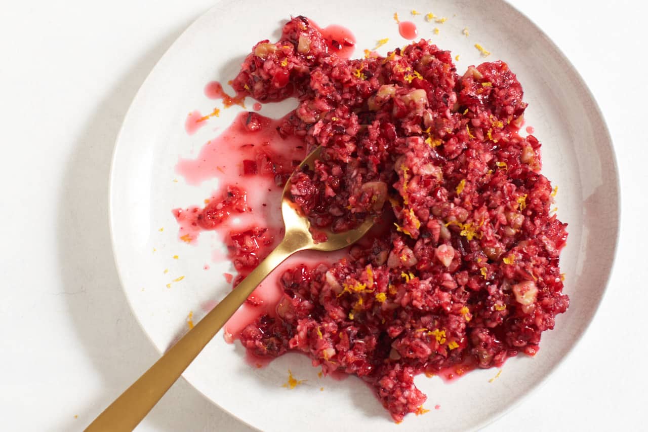 A plate of fresh cranberry orange relish with orange zest on top and a gold spoon on the left side.