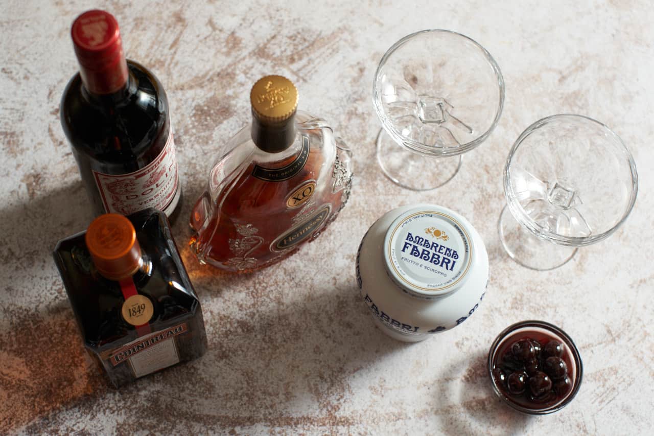 Bottles of cognac, cointreau, and sweet vermouth alongside two coupe glasses, and a jar and bowl of amarena cherries.
