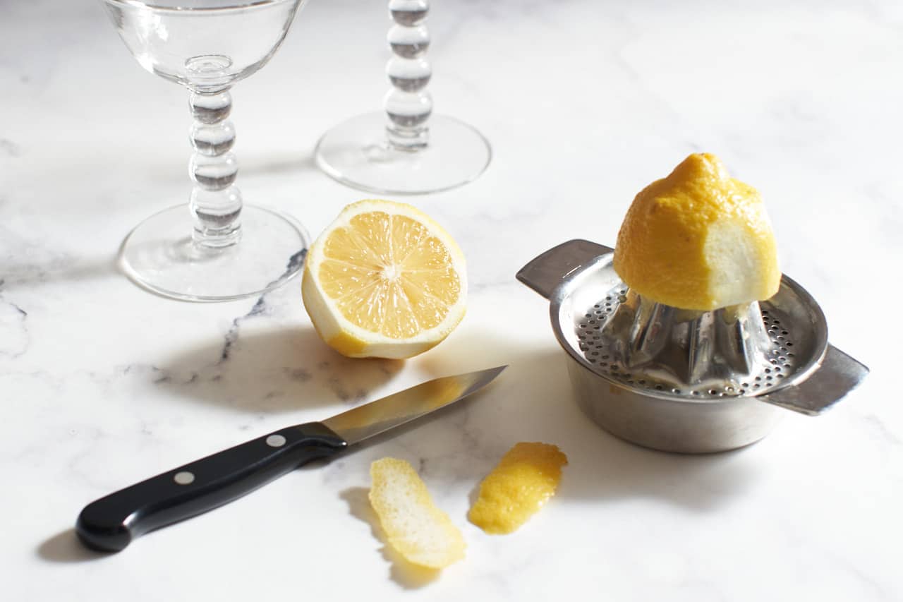 Two coupe glasses next to a halved lemon, two lemon peels, a paring knife, and a lemon on a juicer.