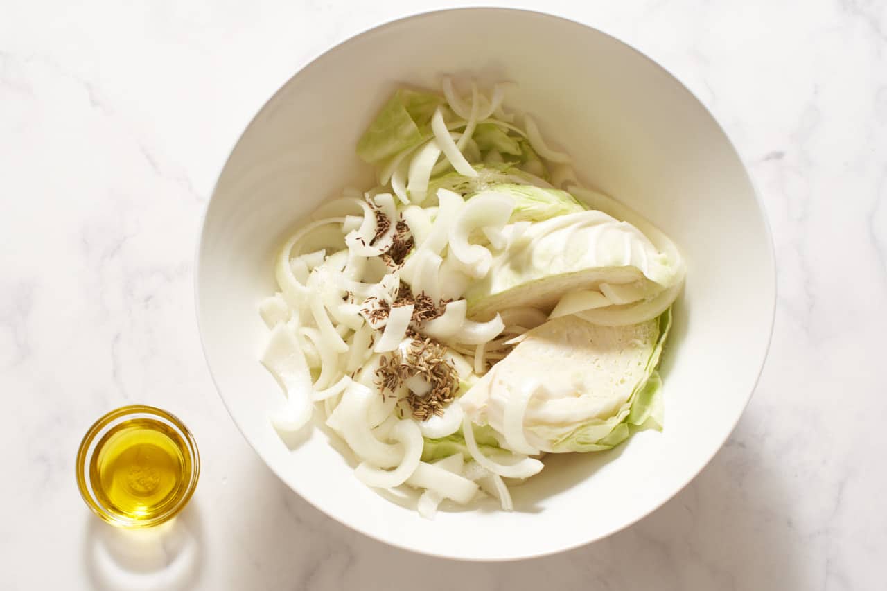 A small bowl of olive oil next to a bowl of cabbage wedges and sliced onions with spices.