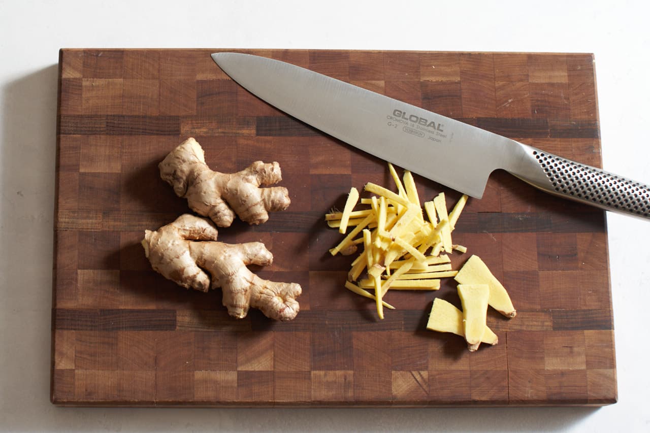 A knife on a cutting board with stalks of ginger, and ginger sliced into matchsticks.