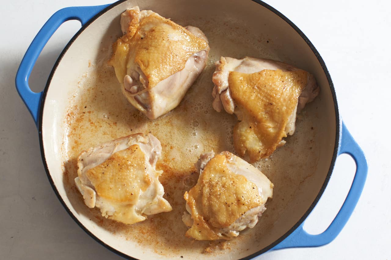 Four lightly golden brown chicken thighs in a casserole pan with blue handles.
