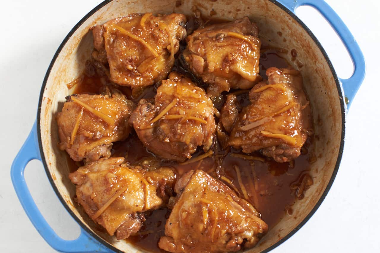 Vietnamese caramel chicken after the sauce has been reduced in a pan with blue handles. Sliced ginger and sauce are clinging to the chicken.