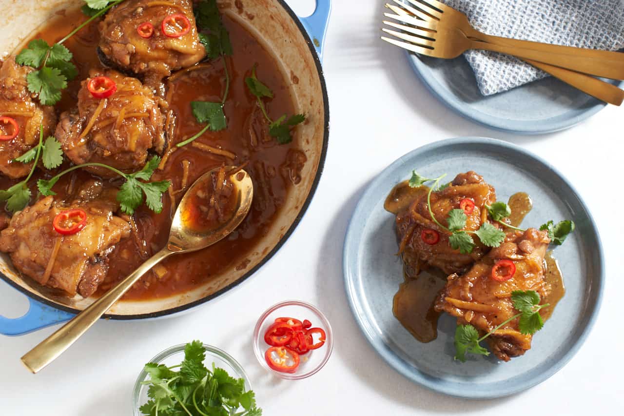 A pan of Vietnamese caramel chicken with a gold spoon in it is on the left. Two chicken thighs covered in sauce are on a plate to the right. Gold forks are in the upper right, small bowls of garnishes are in the lower left.