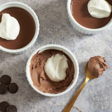 Three chocolate pots de creme with whipped cream on top. Chocolate wafers are on the left, a spoon with chocolate on it is on the right.