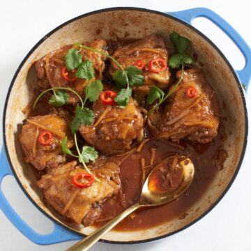 Vietnamese caramel chicken topped with cilantro and red chilis in a pan with blue handles. A gold spoon rests in the pan sauce toward the bottom right.