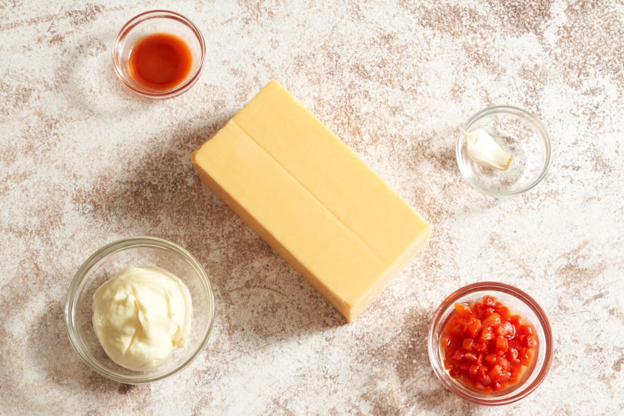 A block of sharp cheddar cheese surrounded by small glass bowls of pimentos, mayonnaise, hot sauce, and garlic.