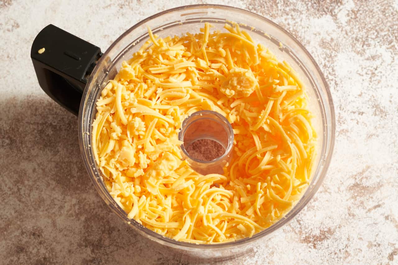 The bowl of a food processor filled with grated cheddar cheese.
