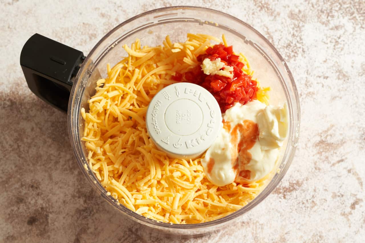The bowl of a food processor filled with grated cheddar cheese, pimentos, mayonnaise, and grated garlic.