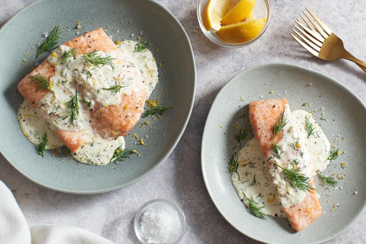 Two green plates with salmon topped with creamy dill sauce and garnished with dill and lemon zest. A small bowl of salt is at the bottom, a bowl of lemon slices and two gold forks are in the top right.