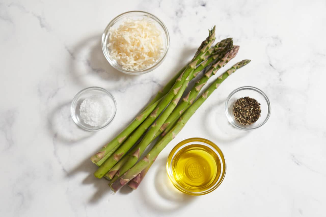 A bunch of fresh asparagus surrounded by small glass bowls of salt, pepper, olive oil, and parmesan cheese.