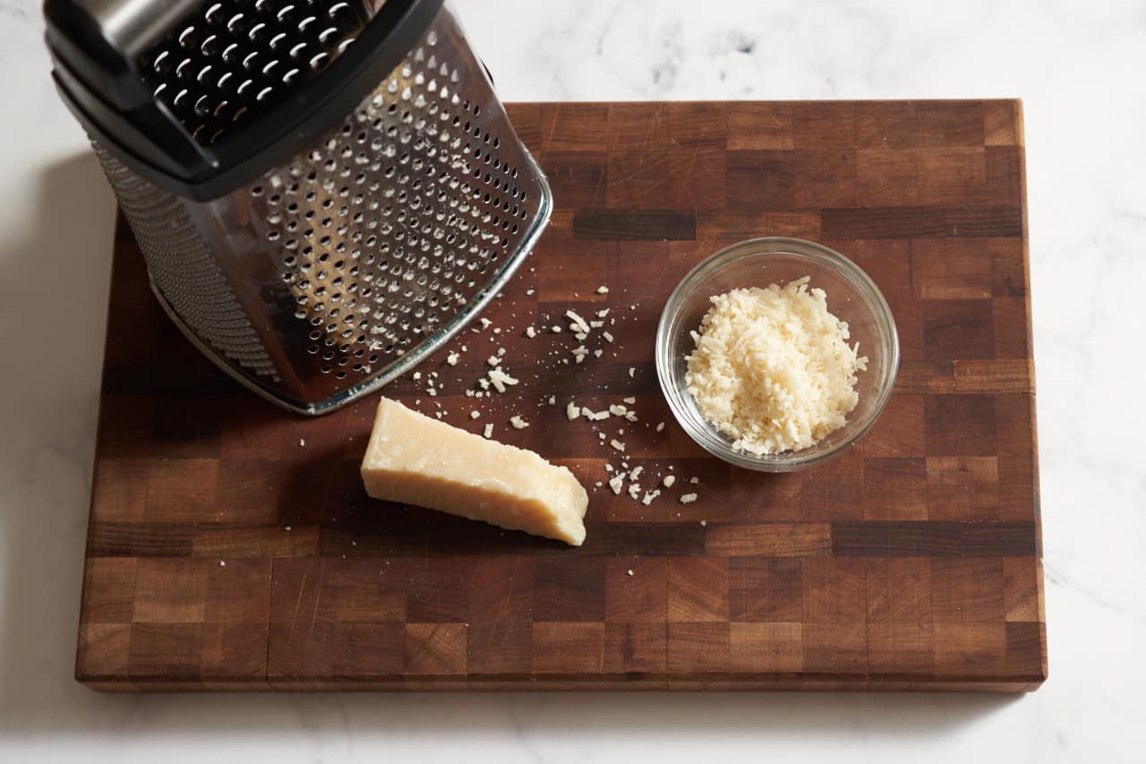 A box grater on a cutting board with a piece of parmesan and grated parmesan cheese.