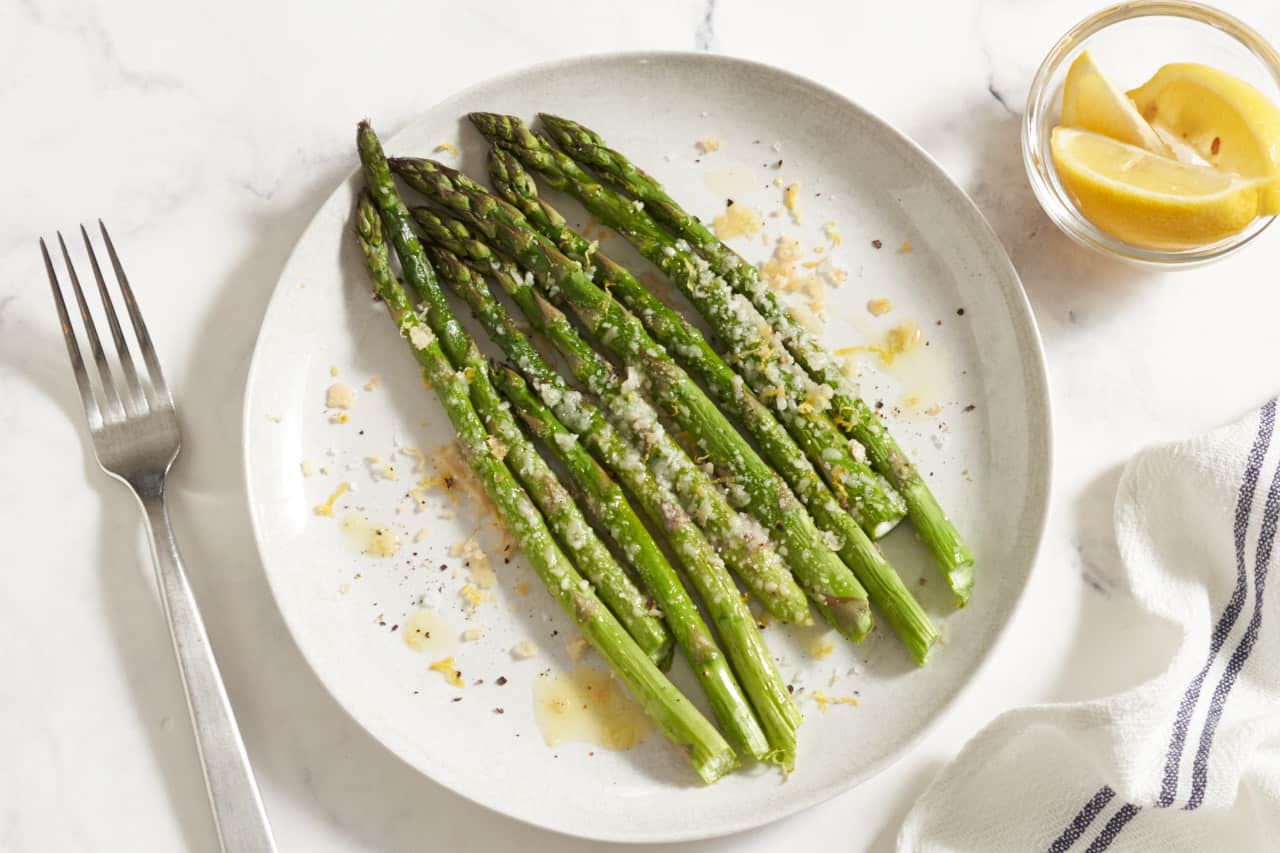Baked asparagus with parmesan cheese on a white plate. A fork is to the left, a small bowl of lemon wedges and a blue and white striped napkin are on the right.