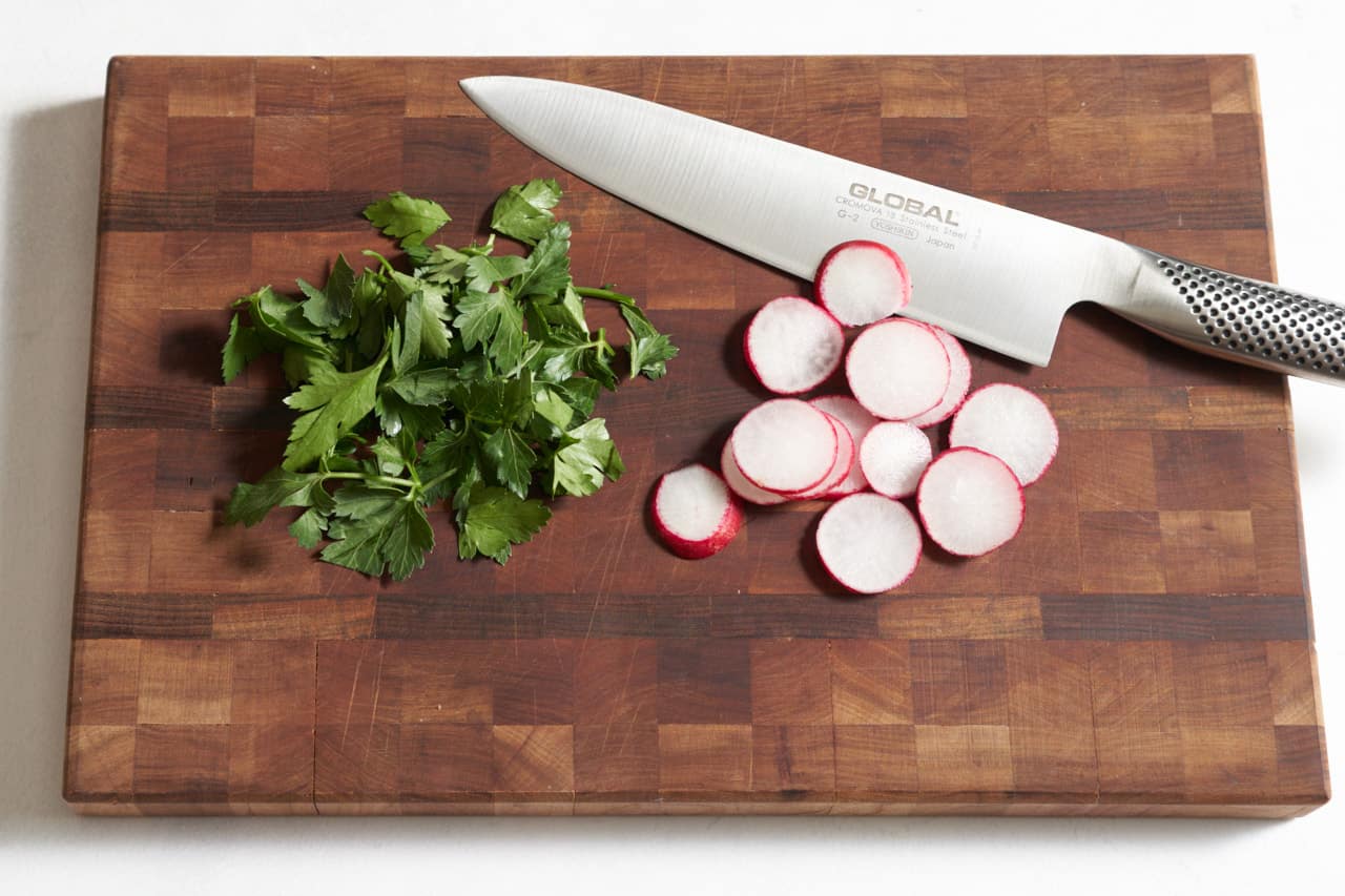 A knife on a cutting board with chopped parsley and sliced radishes.